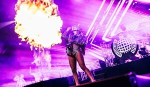 An energetic performer on stage amidst vibrant purple lights, with a dramatic fire explosion lighting up the atmosphere.