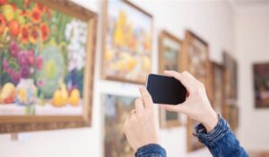A person takes a photo of vibrant artwork on display in a gallery, showcasing colourful paintings with fruit and flowers.
