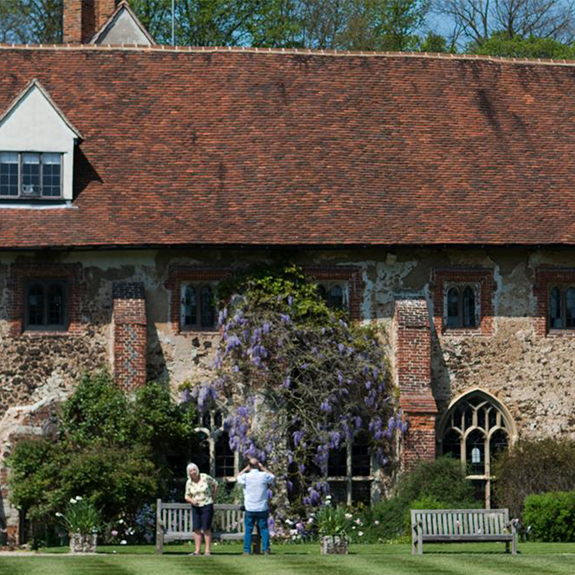 A couple admires a picturesque old building covered in wisteria, surrounded by lush greenery and benches on a sunny day.