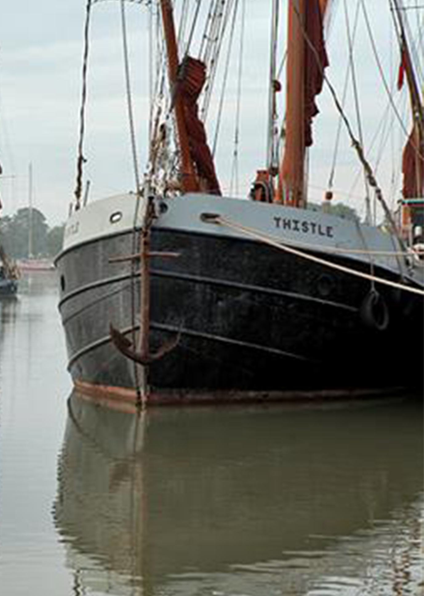 A historic wooden ship named "Thistle" moored calmly in a reflective harbour, with its prominent anchor visible at the bow.