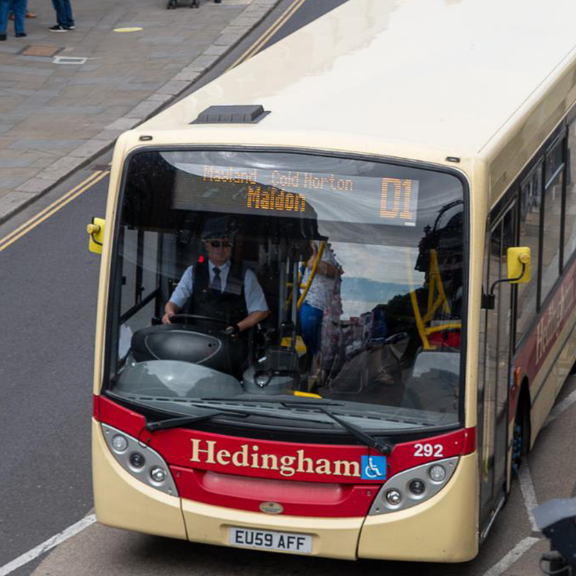 A Hedingham bus, route D1 to Maldon, drives through a city street, with passengers and shops visible in the background.
