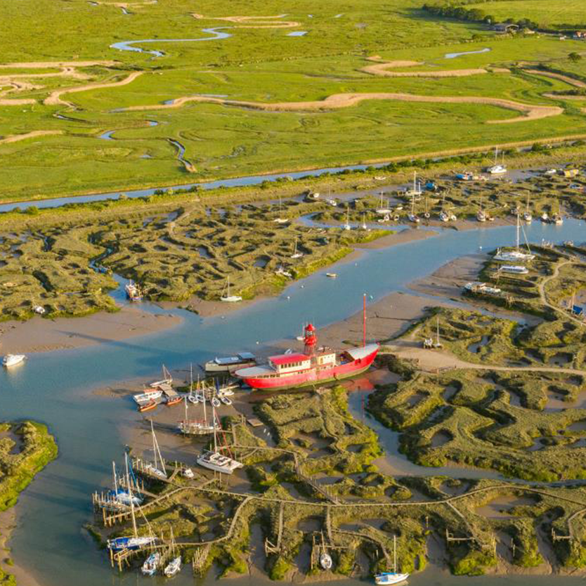 Aerial view of a winding river meandering through green marshlands, featuring a red boat and various other vessels docked in the water.