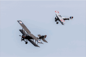 Two vintage biplanes perform aerial manoeuvres against a pale blue sky, showcasing their distinct military insignias.