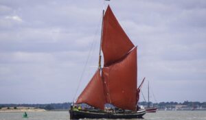 A traditional sailing boat with large red sails navigates through calm waters under a cloudy sky, surrounded by other vessels.