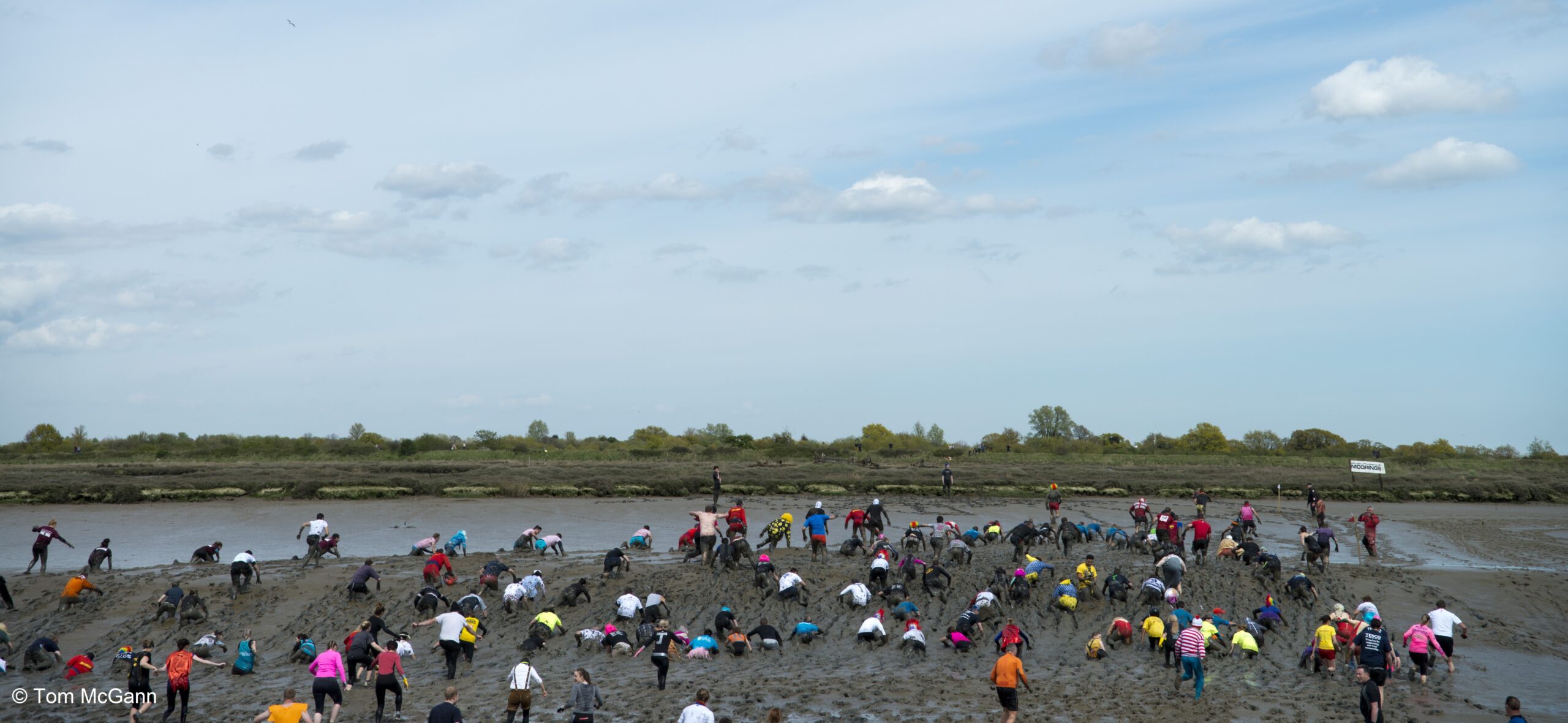 An outdoor, eye-level shot shows a crowd of people in colorful clothes crawling through deep mud in a wide, flat, muddy expanse under a bright sky with scattered clouds. The mud is thick and brown, covering the participants from head to toe as they move forward. In the background, a line of green bushes and trees marks the horizon. The overall impression is one of a challenging and messy, yet seemingly fun, event.