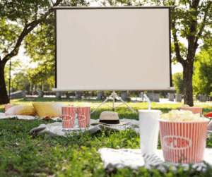 A cinema screen set up in the park with picnic blankets laid on the grass with popcorn and drinks on