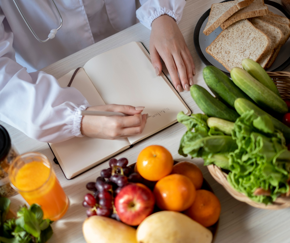 A person writing in a notebook. The word 'Nutrition' on the page. The note book surrounded by fresh fruit and vegetables on the worksurface.
