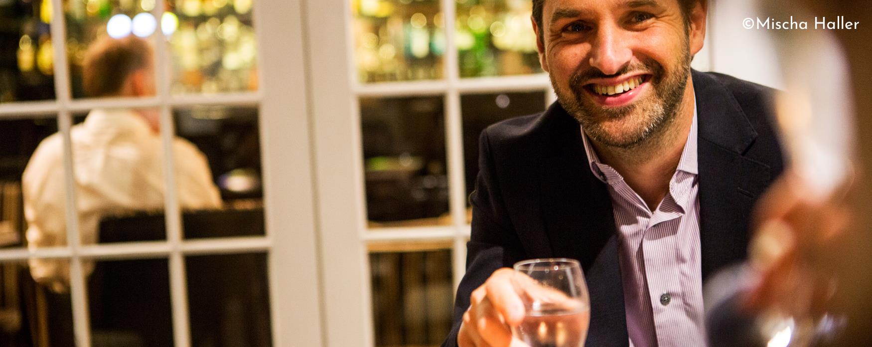 Man in a suit smiling at a dining table with a glass of white wine in his hand