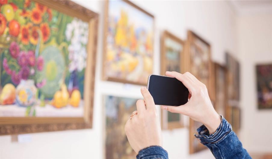 Here is an alt text description for the image:

A person in a denim jacket takes a photo of a painting in a gallery with their smartphone.

Additional