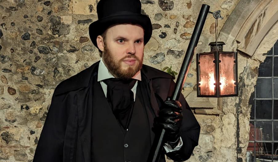 A person dressed in Victorian-style attire stands against a stone wall, holding a black cane and illuminated by a lantern.